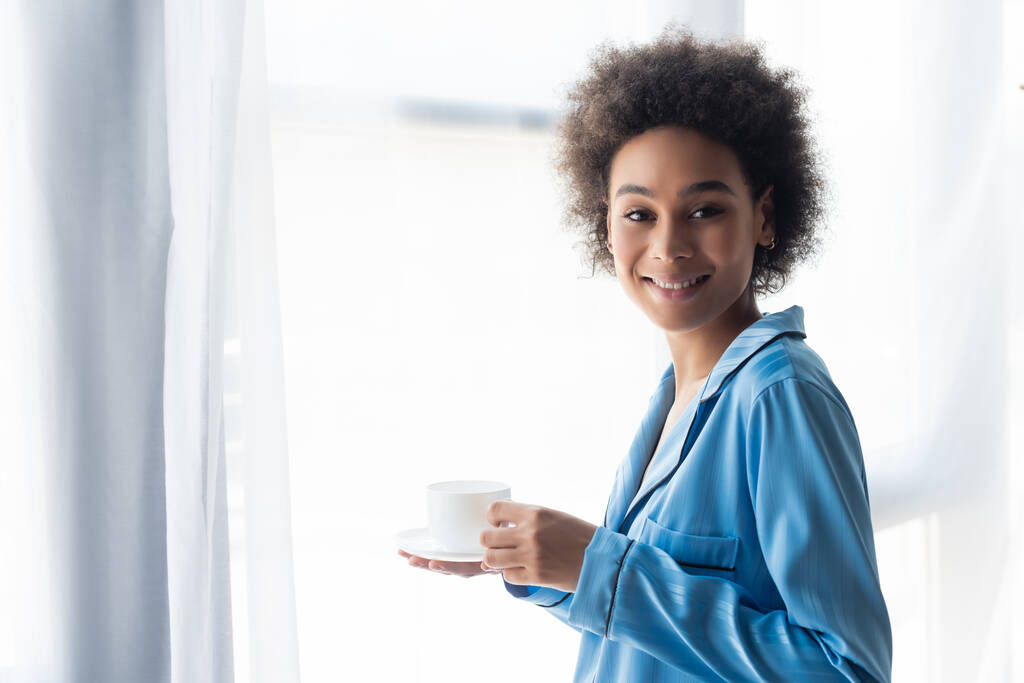 幸せなアフリカ系アメリカ人女性がコーヒーとソーサーを持っていて  - 写真・画像
