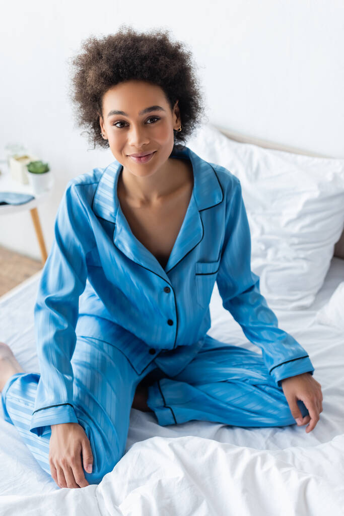 magas szög kilátás pozitív és fiatal afro-amerikai nő pizsamában ül az ágyban - Fotó, kép