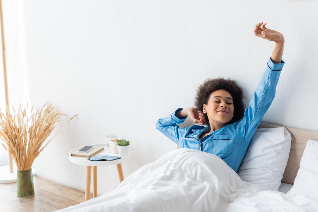 pleased african american woman in pajamas stretching on bed - Photo, Image