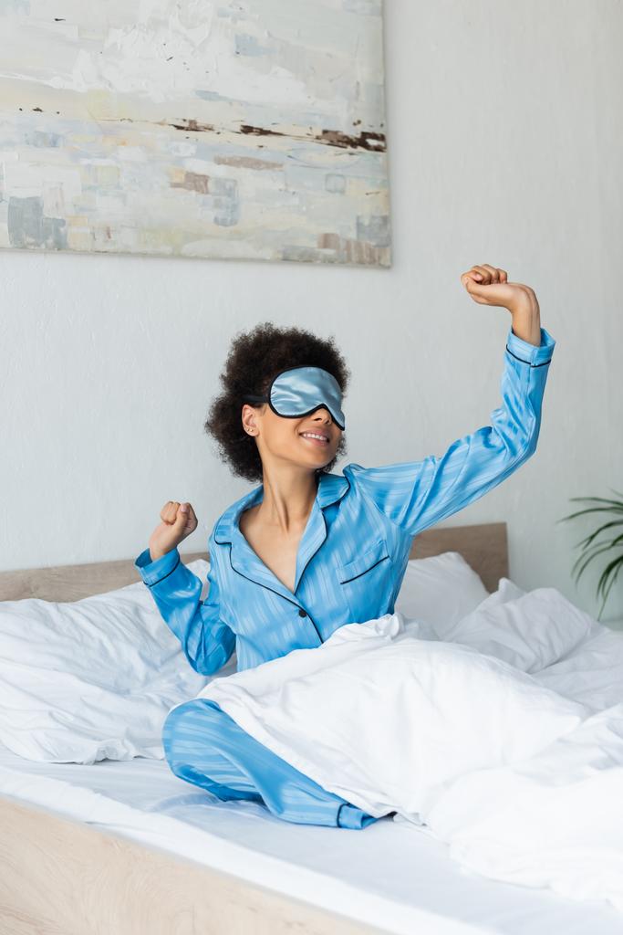 tyytyväinen afrikkalainen amerikkalainen nainen pyjamassa ja nukkuva naamio venyttely sängyssä - Valokuva, kuva