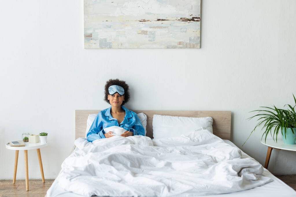 vrolijk Afrikaans amerikaanse vrouw in pyjama en slaapmasker met kopje koffie in bed - Foto, afbeelding