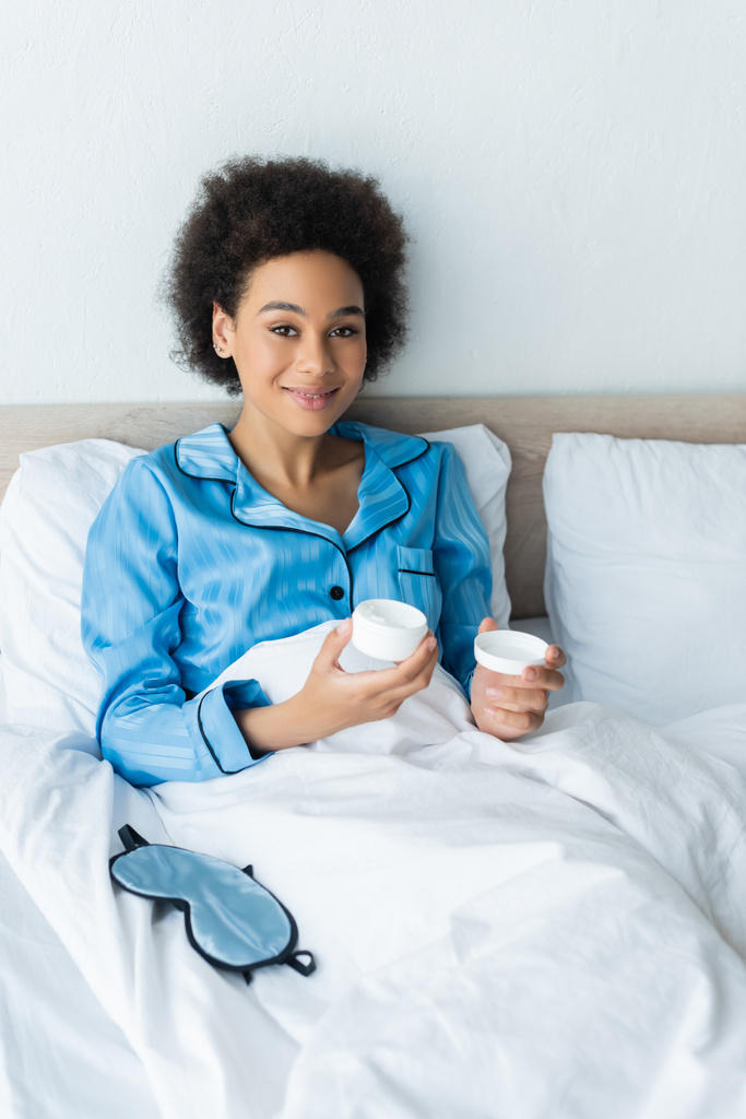 陽気なアフリカ系アメリカ人女性ですパジャマを着て容器に化粧品クリームをベッドで  - 写真・画像