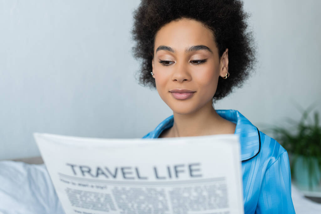 Pijamalı Afrikalı Amerikalı kadın yatak odasında bulanık seyahat gazetesi okuyor.  - Fotoğraf, Görsel