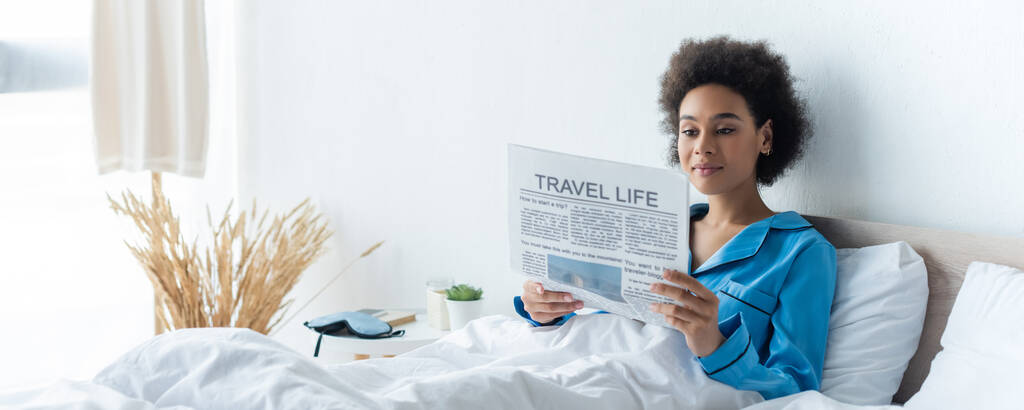 young african american woman in pajamas reading travel life newspaper in bed, banner - Photo, Image