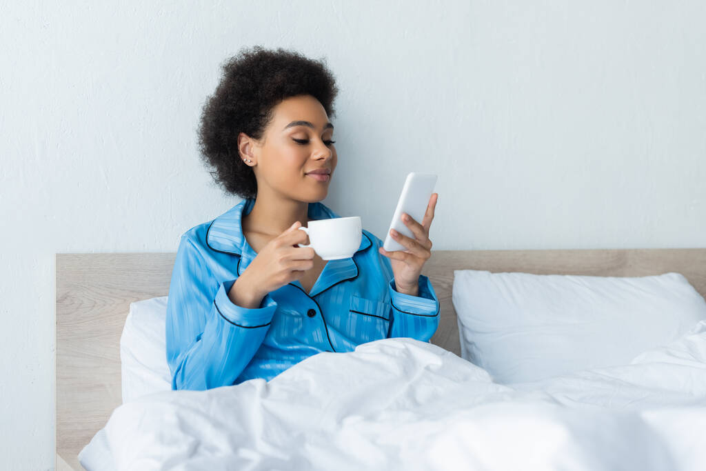 glimlachen Afrikaans amerikaanse vrouw in pyjama met behulp van smartphone en het vasthouden van kopje koffie in bed - Foto, afbeelding