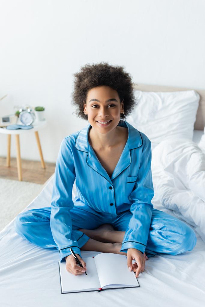 gioiosa donna afroamericana in pigiama seduta sul letto e che scrive sul taccuino - Foto, immagini