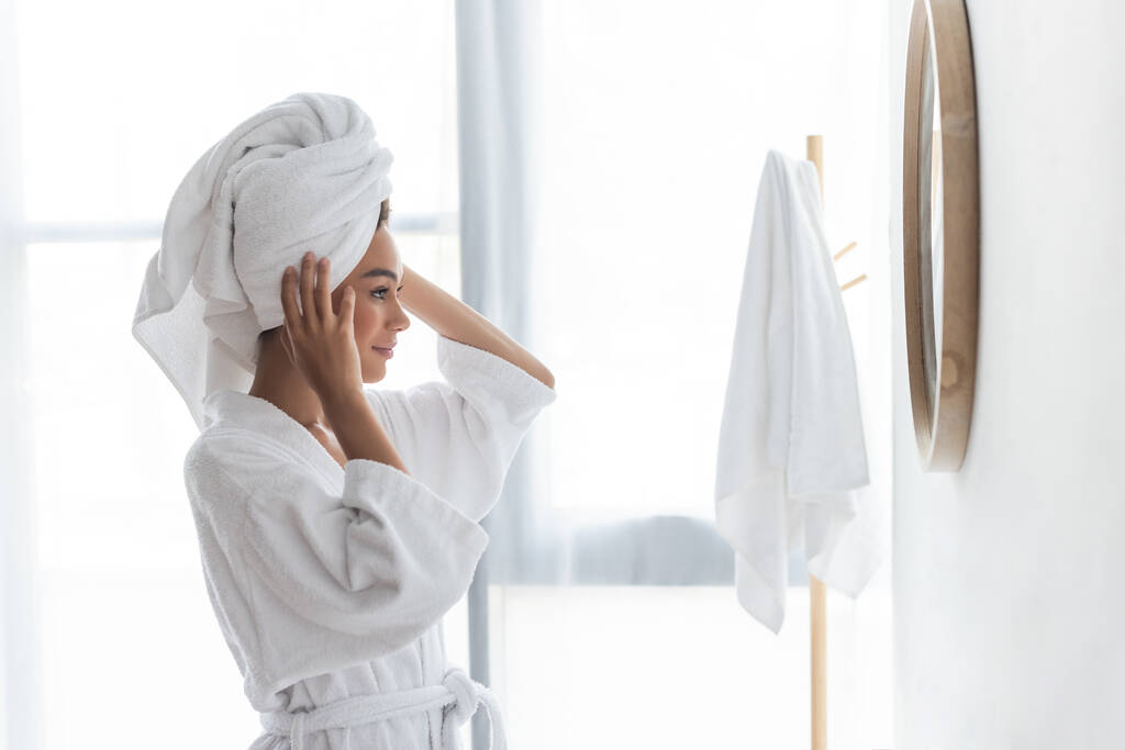 gioiosa donna afro-americana che regola l'asciugamano e guarda lo specchio in bagno - Foto, immagini