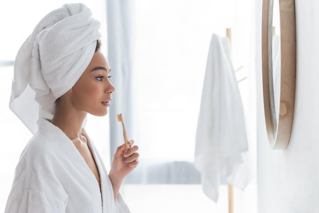 boční pohled na africké americké ženy při pohledu na zrcadlo a držení zubního kartáčku v koupelně - Fotografie, Obrázek