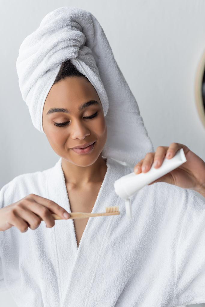 χαρούμενη Αφροαμερικανή γυναίκα με μπουρνούζι και πετσέτα κρατώντας οδοντόκρεμα και οδοντόβουρτσα στο μπάνιο  - Φωτογραφία, εικόνα