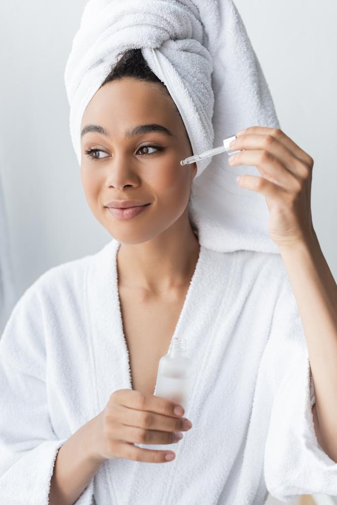 mujer afroamericana complacida en bata blanca sosteniendo la botella y aplicando suero en el baño  - Foto, imagen