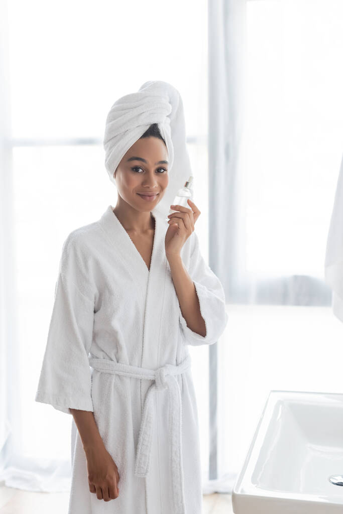 pleased african american woman in white towel and bathrobe holding bottle with serum  - Photo, Image