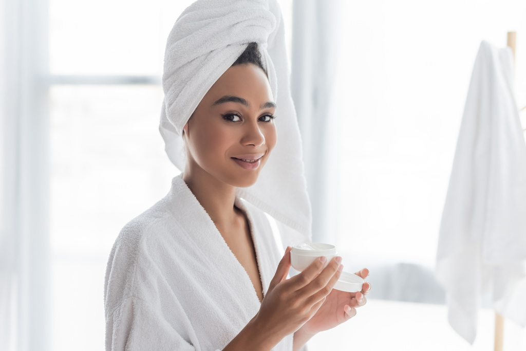 усміхнена афроамериканська жінка в халаті тримає контейнер з кремом для обличчя у ванній
  - Фото, зображення