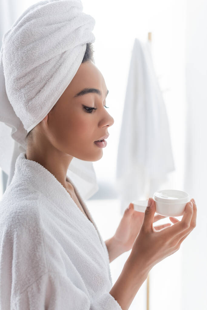 african american woman in bathrobe looking at container with face cream in bathroom  - Photo, Image