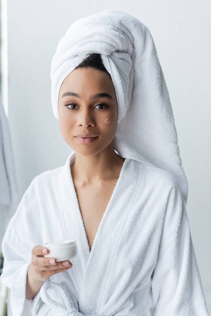 mujer afroamericana en bata de baño sosteniendo contenedor con crema facial  - Foto, Imagen