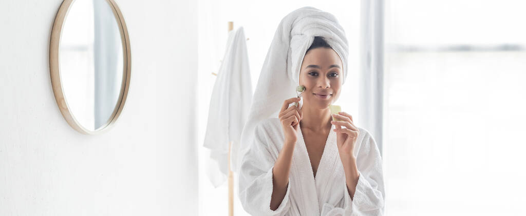 happy african american woman in bathrobe massaging face with jade roller and face scraper, banner - Photo, Image