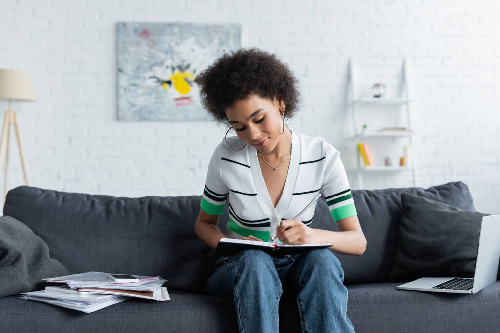 curly african american woman writing in notebook near laptop on couch - Photo, Image