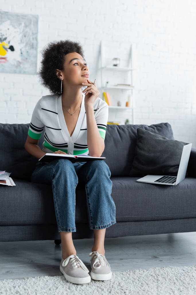 nachdenkliche Afroamerikanerin sitzt mit Notizbuch neben Laptop auf Couch - Foto, Bild