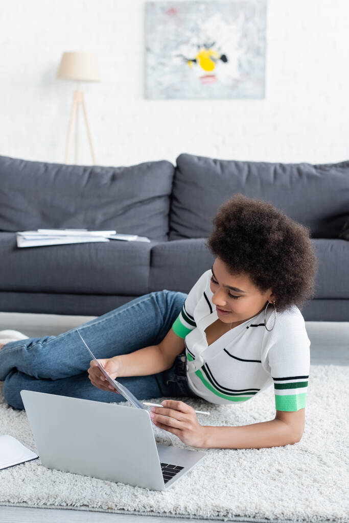 allegro afroamericano freelance guardando il documento vicino al computer portatile mentre sdraiato su tappeto  - Foto, immagini