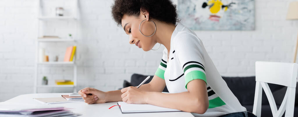 Side view of african american woman writing on notebook near papers at home, banner  - Photo, Image