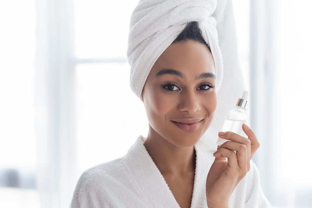 Smiling african american woman in bathrobe holding serum at home  - Photo, Image