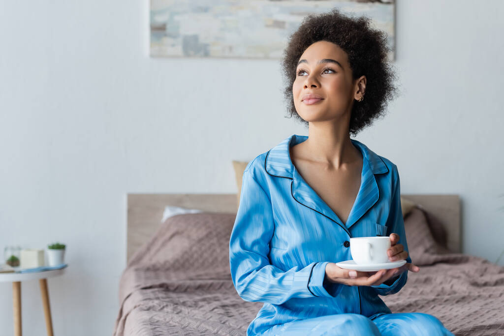 Jeune femme afro-américaine en pyjama tenant une tasse blanche et une soucoupe dans la chambre  - Photo, image
