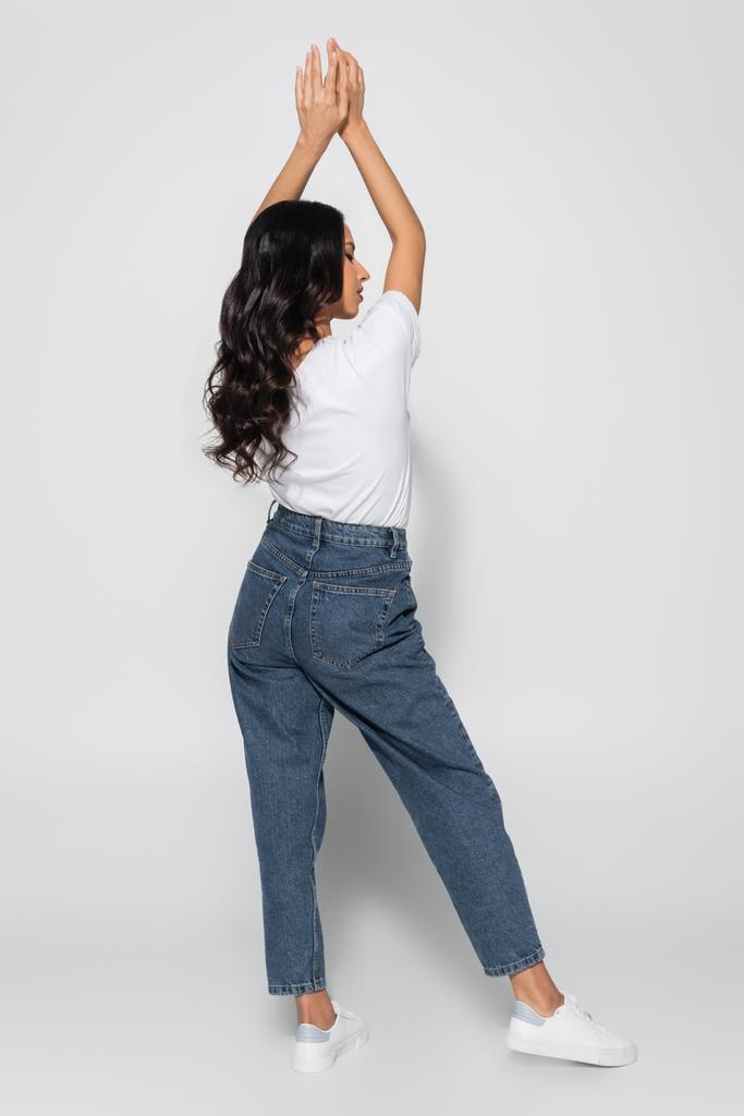 полный вид женщины в белой футболке и джинсах, стоящей с поднятыми руками на сером - Фото, изображение