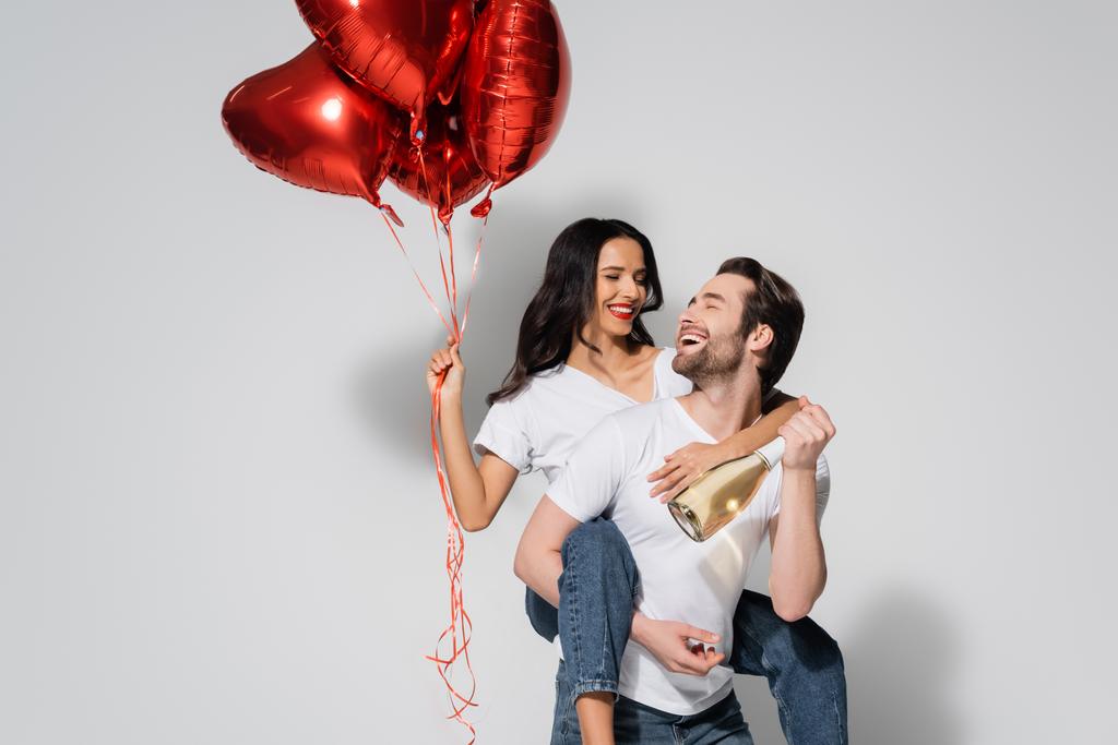alegre hombre con champán botella piggybacking mujer con rojo en forma de corazón globos en gris - Foto, imagen