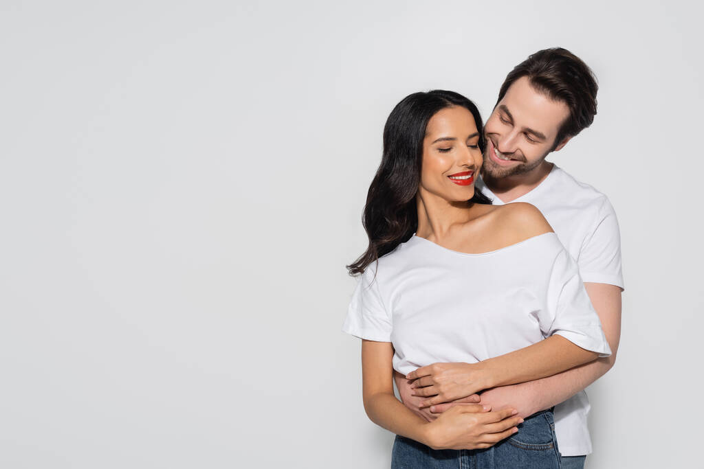 fröhlicher Mann umarmt brünette Frau in weißem T-Shirt isoliert auf grau - Foto, Bild