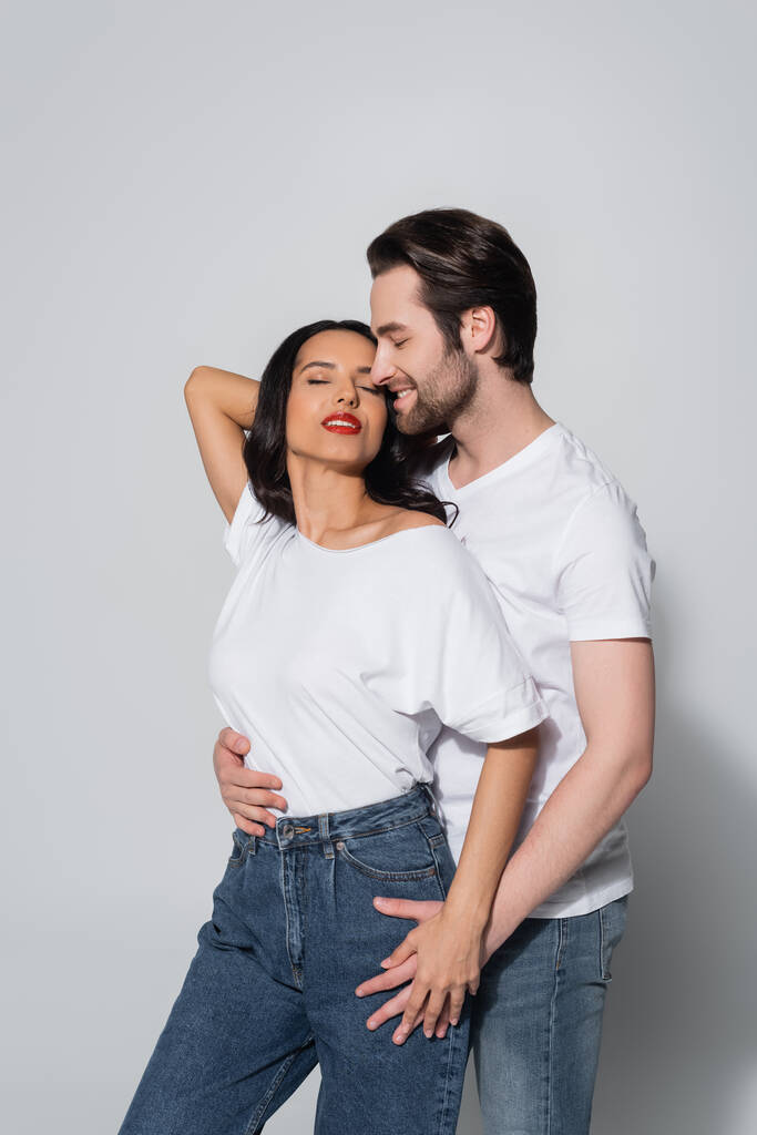 Улыбающийся мужчина обнимает соблазнительную женщину, стоящую с рукой за головой и закрытыми глазами на сером - Фото, изображение