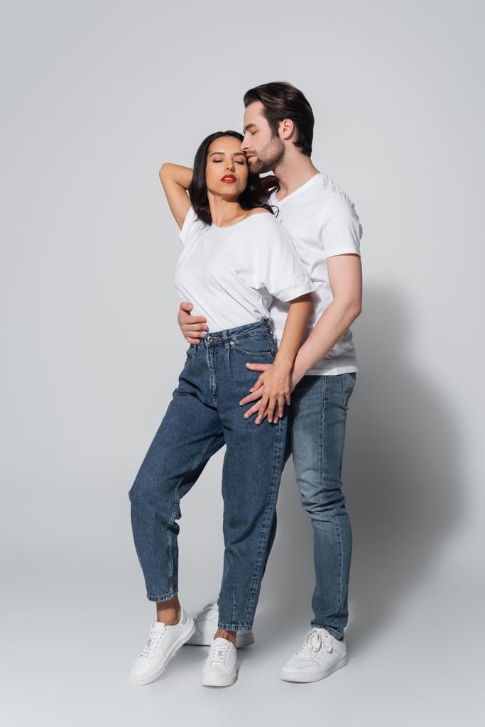 vista completa del hombre con los ojos cerrados abrazando a la mujer seductora en camiseta blanca y jeans en gris - Foto, Imagen