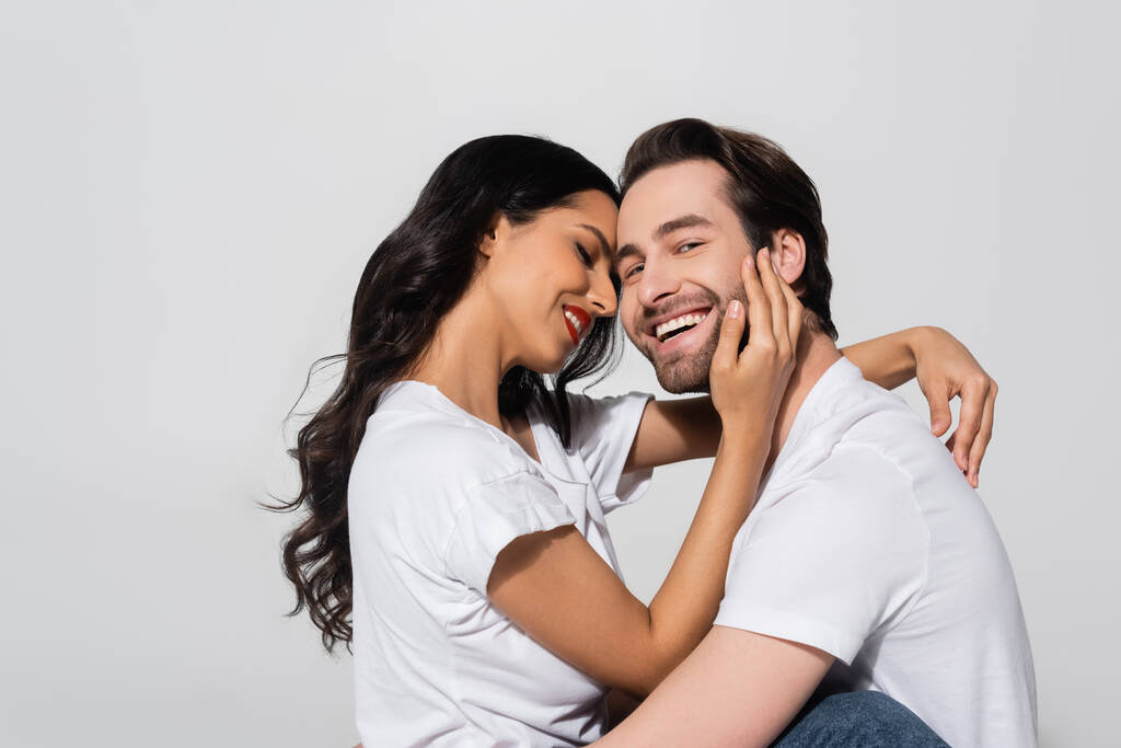 fröhlicher Mann blickt in die Kamera in der Nähe sexy brünette Frau umarmt sein Gesicht isoliert auf grau - Foto, Bild