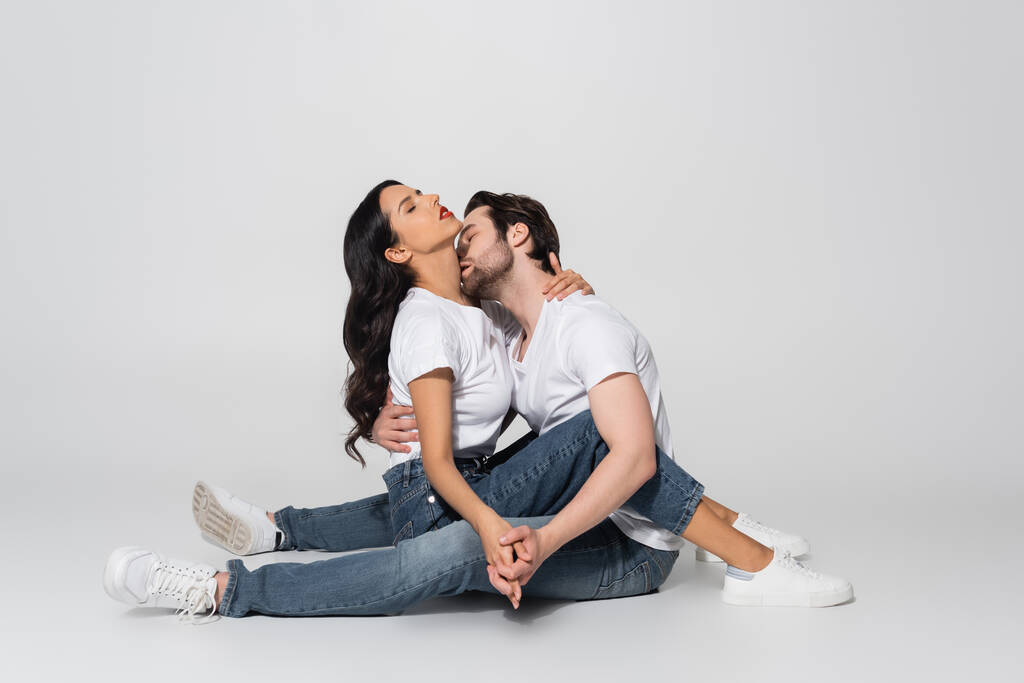 uomo in t-shirt bianca e jeans che si tiene per mano con donna seducente e la bacia mentre è seduta sul grigio - Foto, immagini