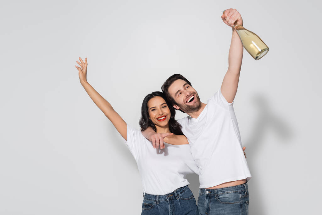 веселый мужчина, держащий бутылку шампанского и указывающий пальцем, обнимая подружку на сером - Фото, изображение