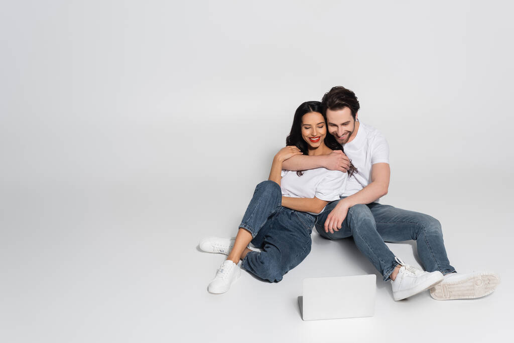 glückliches Paar in lässiger Kleidung beim Ansehen von Film auf Laptop auf grau - Foto, Bild