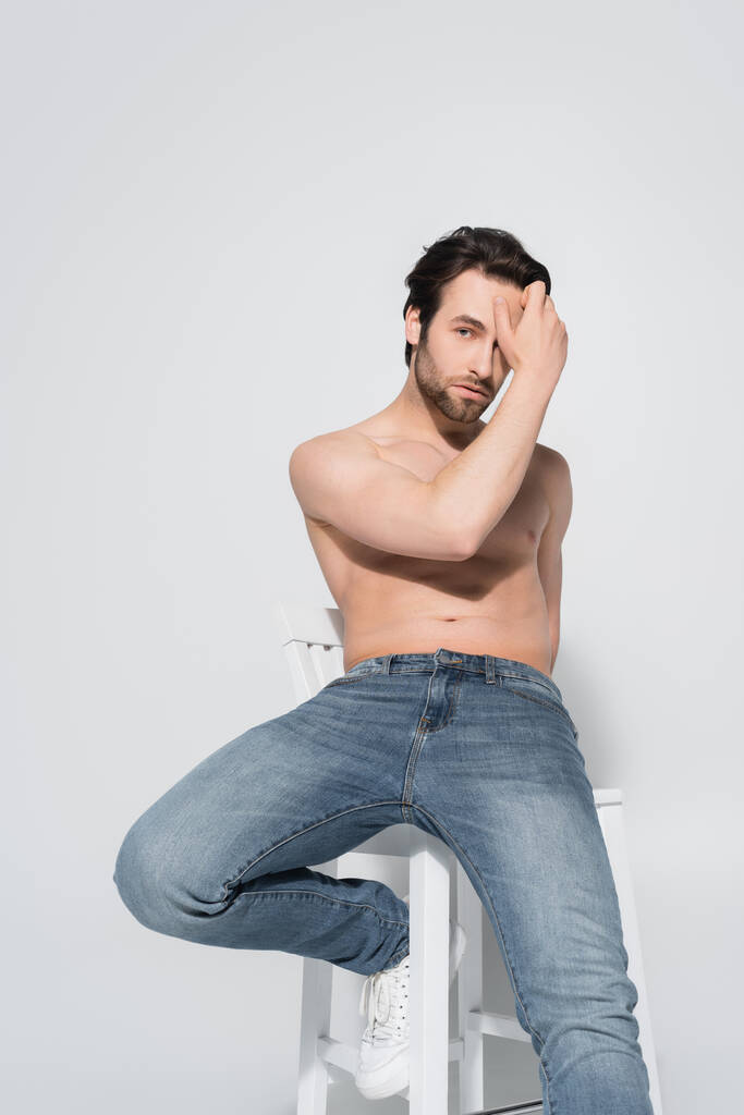 Брюнетка без рубашки мужчина в джинсах смотрит в камеру, держа руку возле лица на сером - Фото, изображение