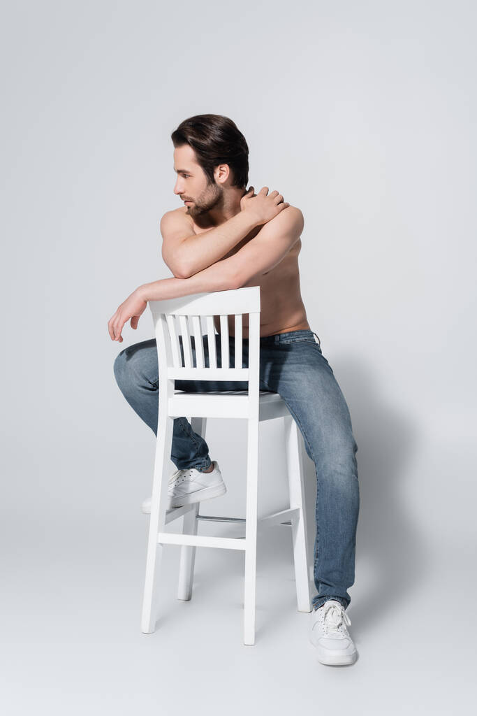 白い椅子に座ってグレーを見ながらジーンズに身を包んだセクシーな白人男性の完全な長さビュー - 写真・画像