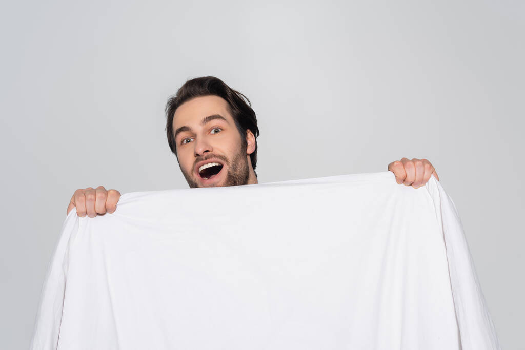 amazed brunette man looking at camera behind white blanket isolated on grey - Photo, Image
