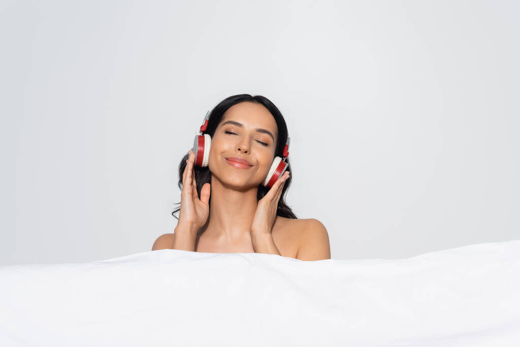 mujer sonriente con los ojos cerrados escuchando música en auriculares detrás de manta blanca aislada en gris - Foto, imagen