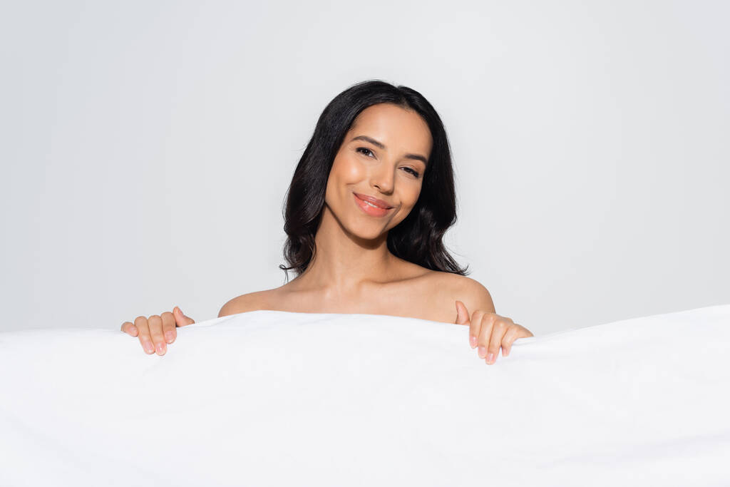 μελαχρινή γυναίκα χαμογελά στην κάμερα, ενώ ποζάρουν πίσω από λευκή κουβέρτα απομονωμένη σε γκρι - Φωτογραφία, εικόνα