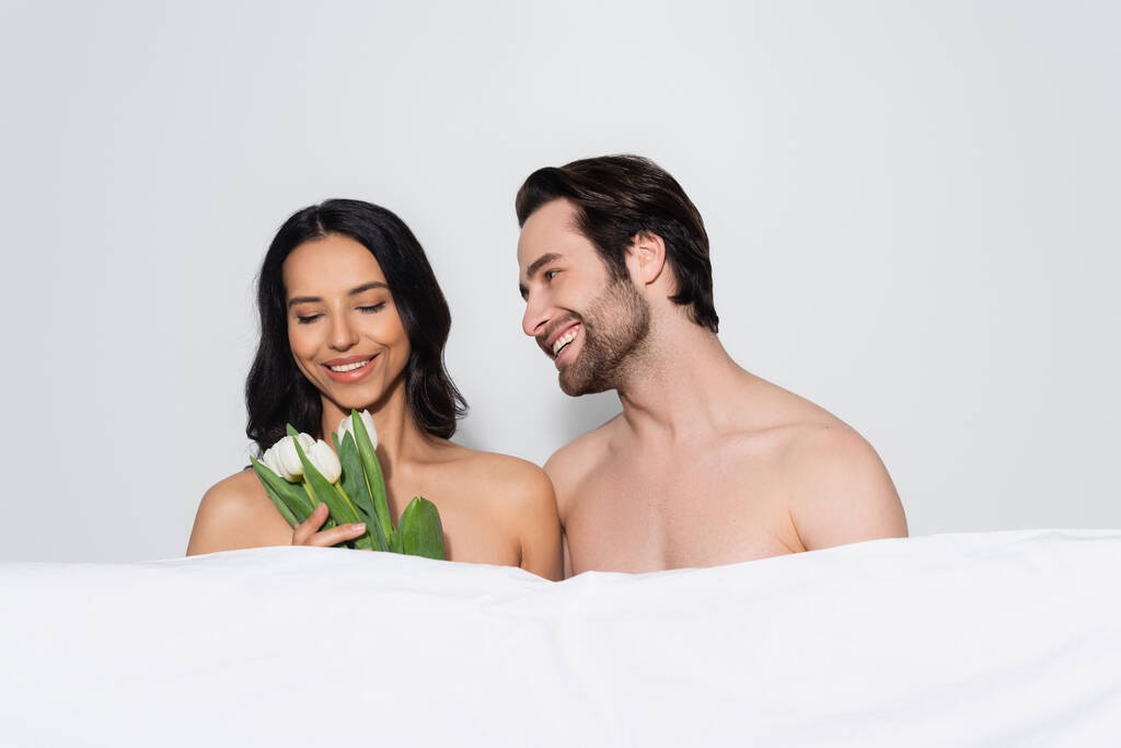χαμογελαστή αισθησιακή γυναίκα που κρατάει τουλίπες κοντά σε γυμνόστηθο άντρα και λευκή κουβέρτα που απομονώνονται σε γκρι - Φωτογραφία, εικόνα