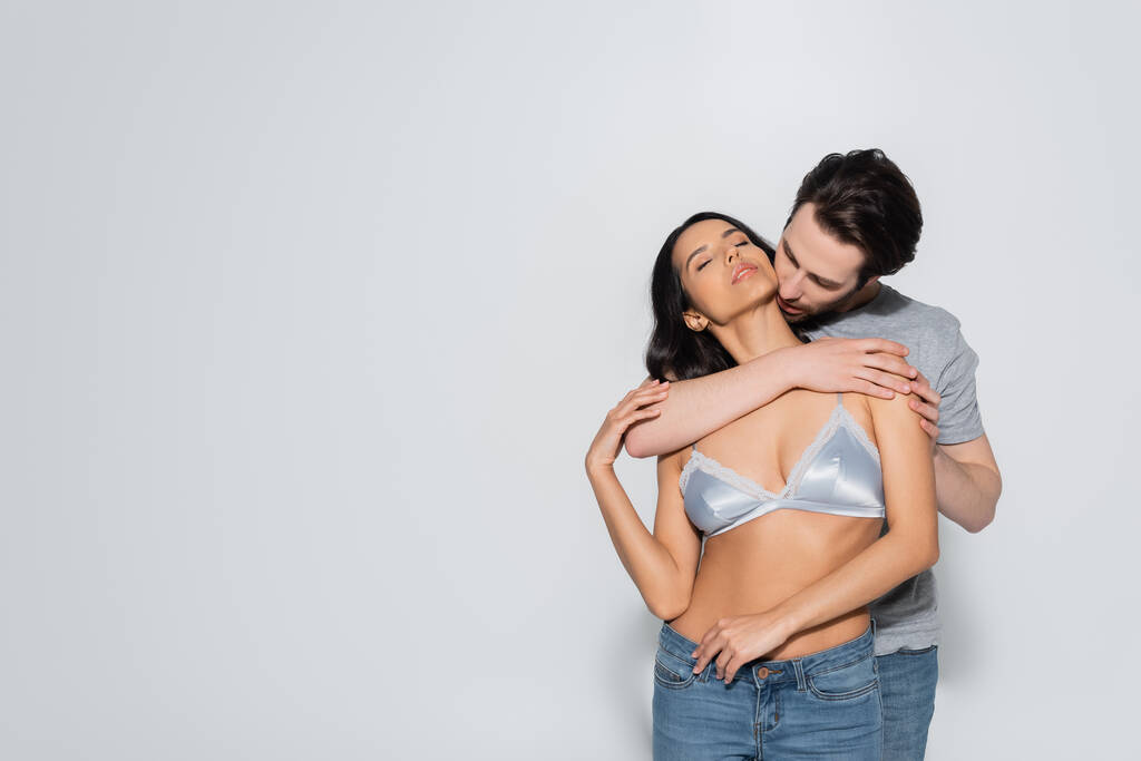 sexy mujer en jeans y sujetador cerca de morena hombre abrazándola en gris - Foto, Imagen