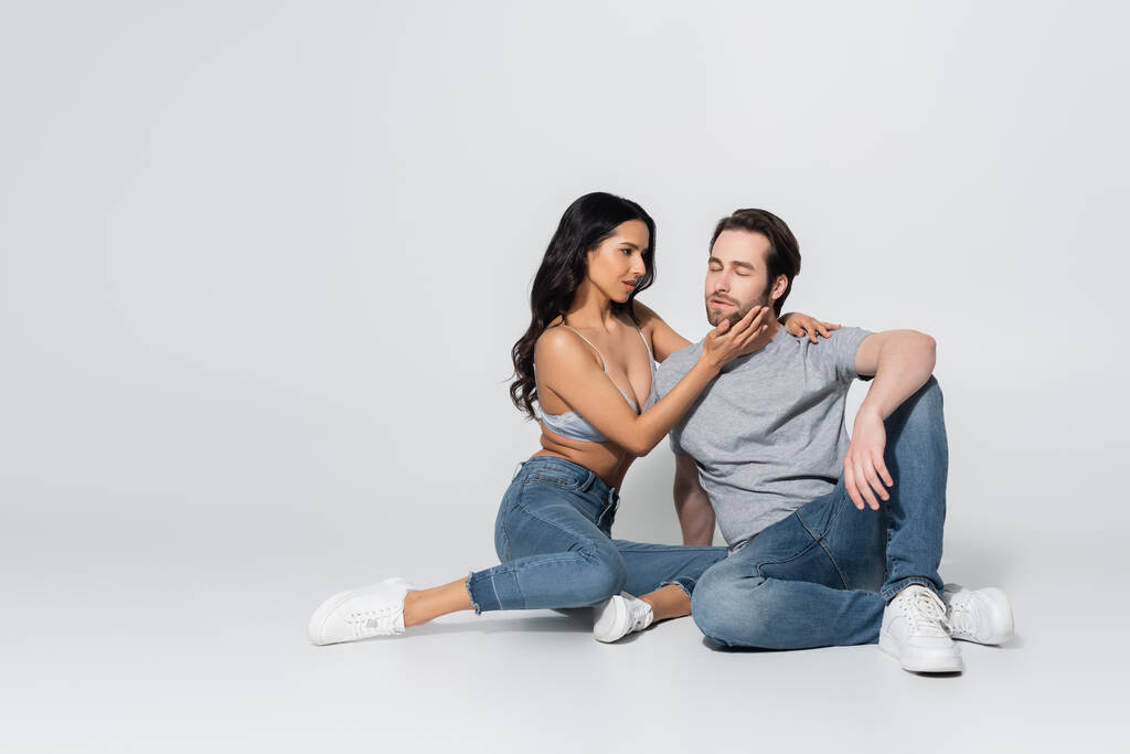 full length view of passionate woman in jeans and bra touching face of young man while sitting on grey - Photo, Image