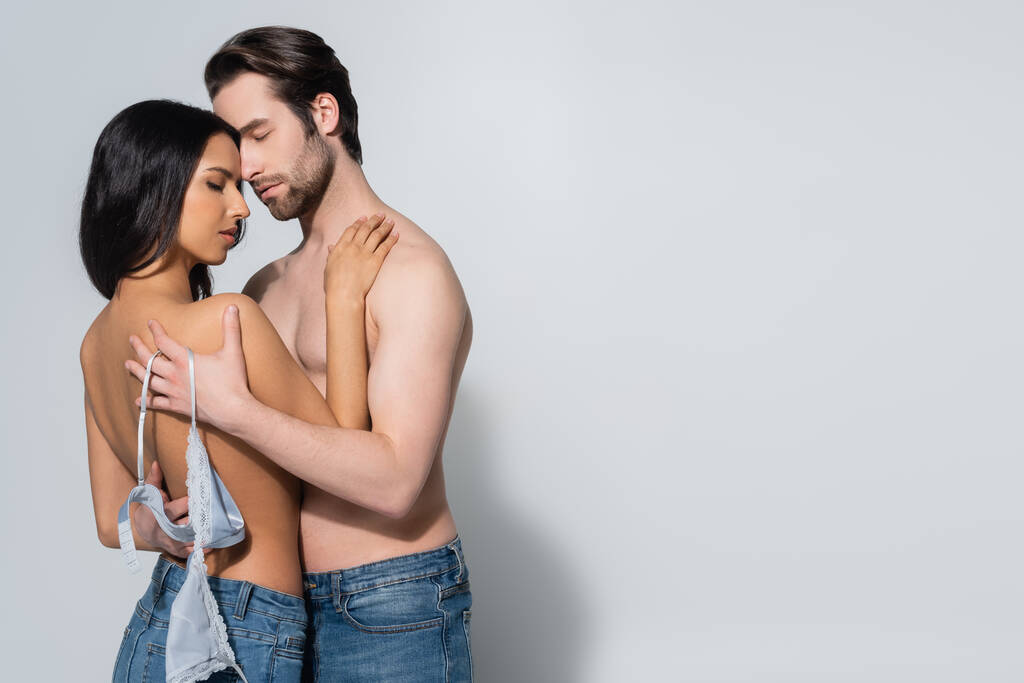 bez koszulki mężczyzna trzymając stanik sexy kobieta w dżinsach obejmując ją na szary  - Zdjęcie, obraz