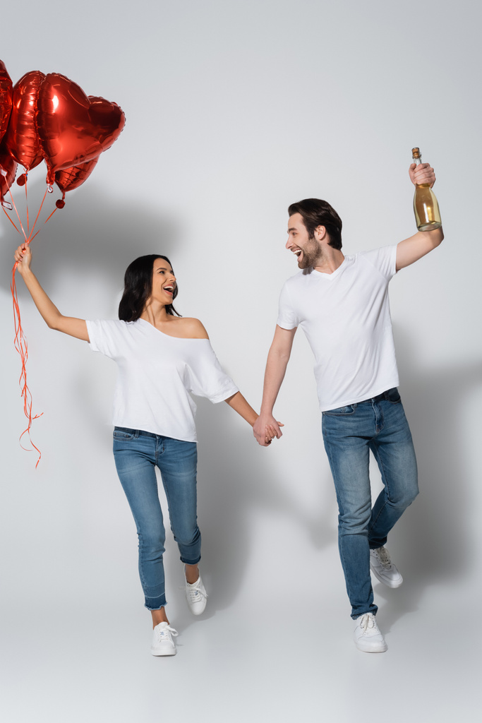 vista completa de pareja feliz cogida de la mano mientras camina con globos rojos en forma de corazón en gris - Foto, imagen