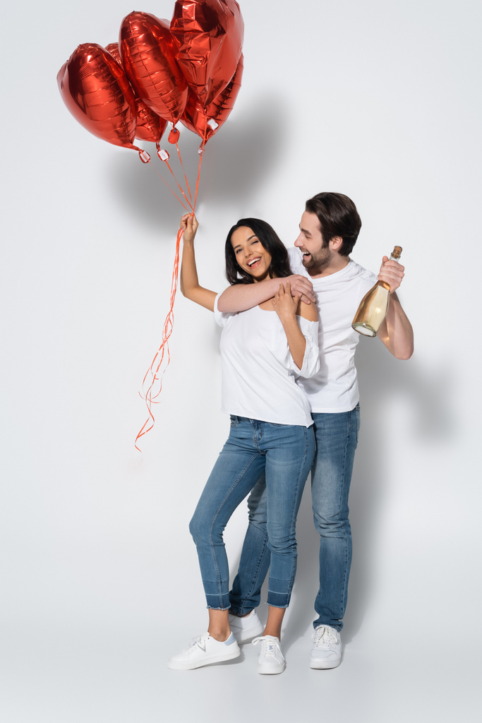 felice uomo con bottiglia di champagne abbracciando fidanzata con palloncini a forma di cuore su grigio - Foto, immagini