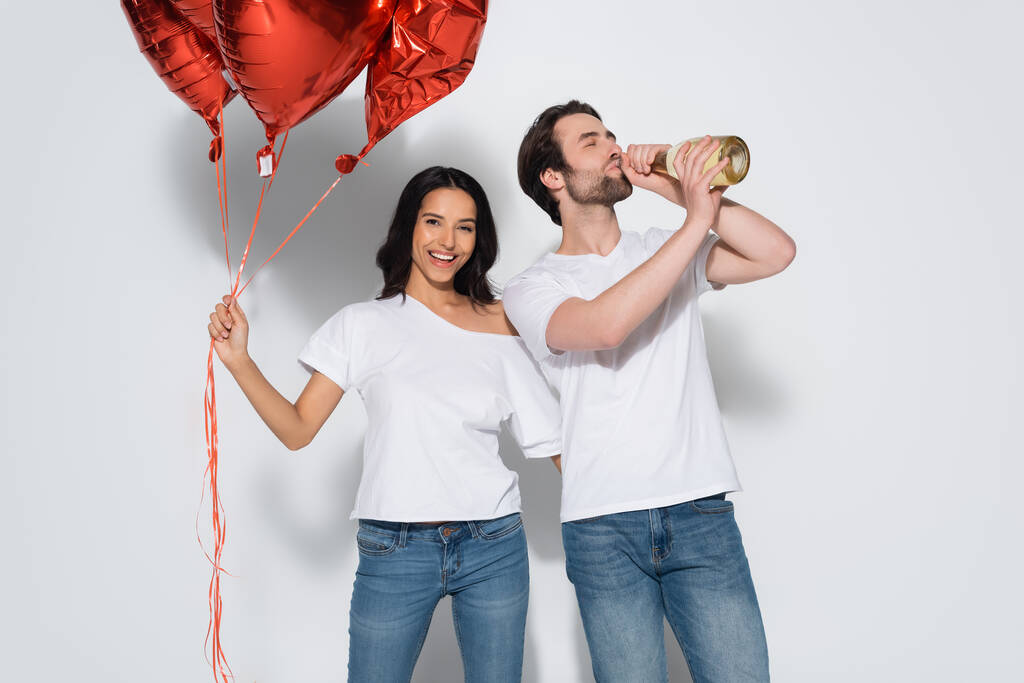 mujer alegre con globos rojos mirando a la cámara cerca de hombre beber champán de botella en gris - Foto, imagen