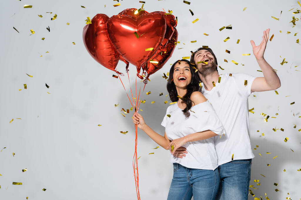 excited couple with heart-shaped balloons laughing under falling confetti on grey - Photo, Image