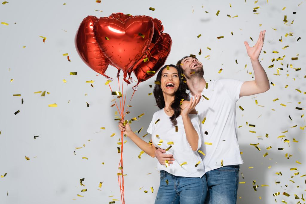 coppia allegra con palloncini a forma di cuore che ridono sotto la caduta di coriandoli sul grigio - Foto, immagini