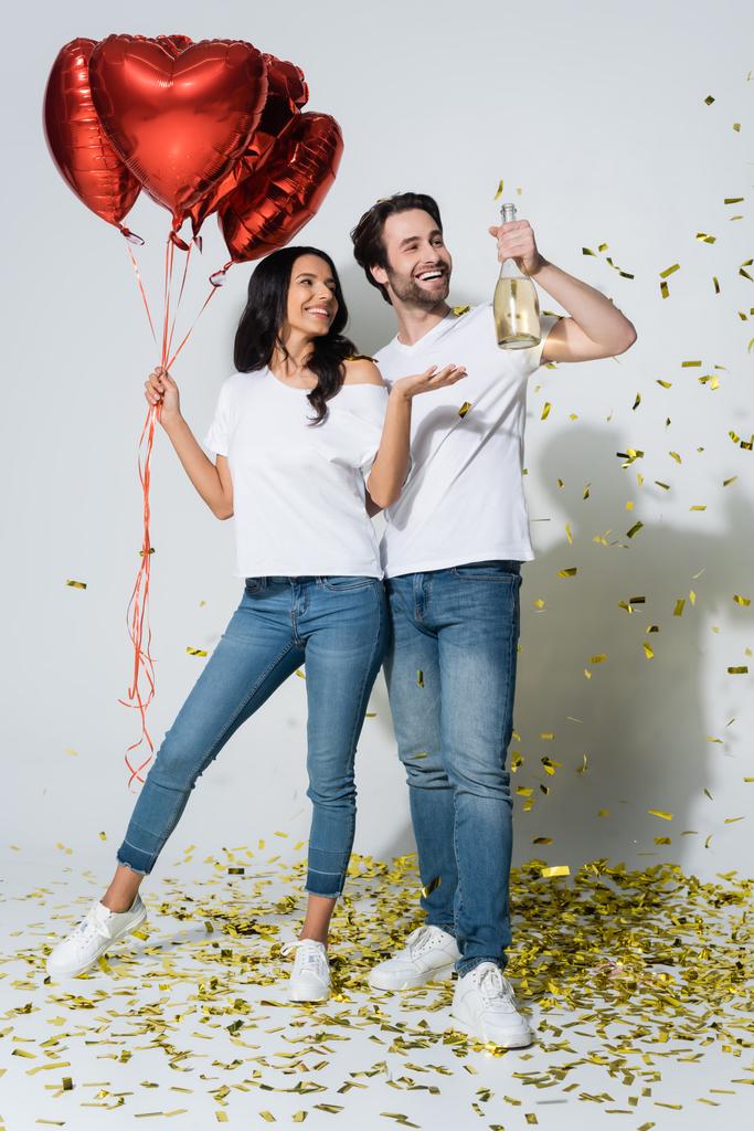 mujer feliz con globos en forma de corazón apuntando a la botella de champán en la mano del novio en gris - Foto, imagen