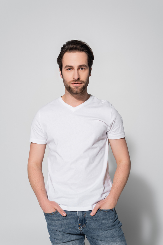 bruna uomo in t-shirt bianca in piedi con le mani in tasche di jeans su grigio - Foto, immagini